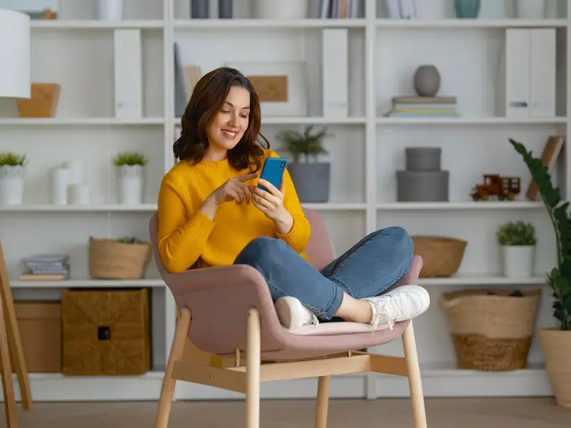 woman sat on chair with her legs crossed, smiling down at her mobile phone